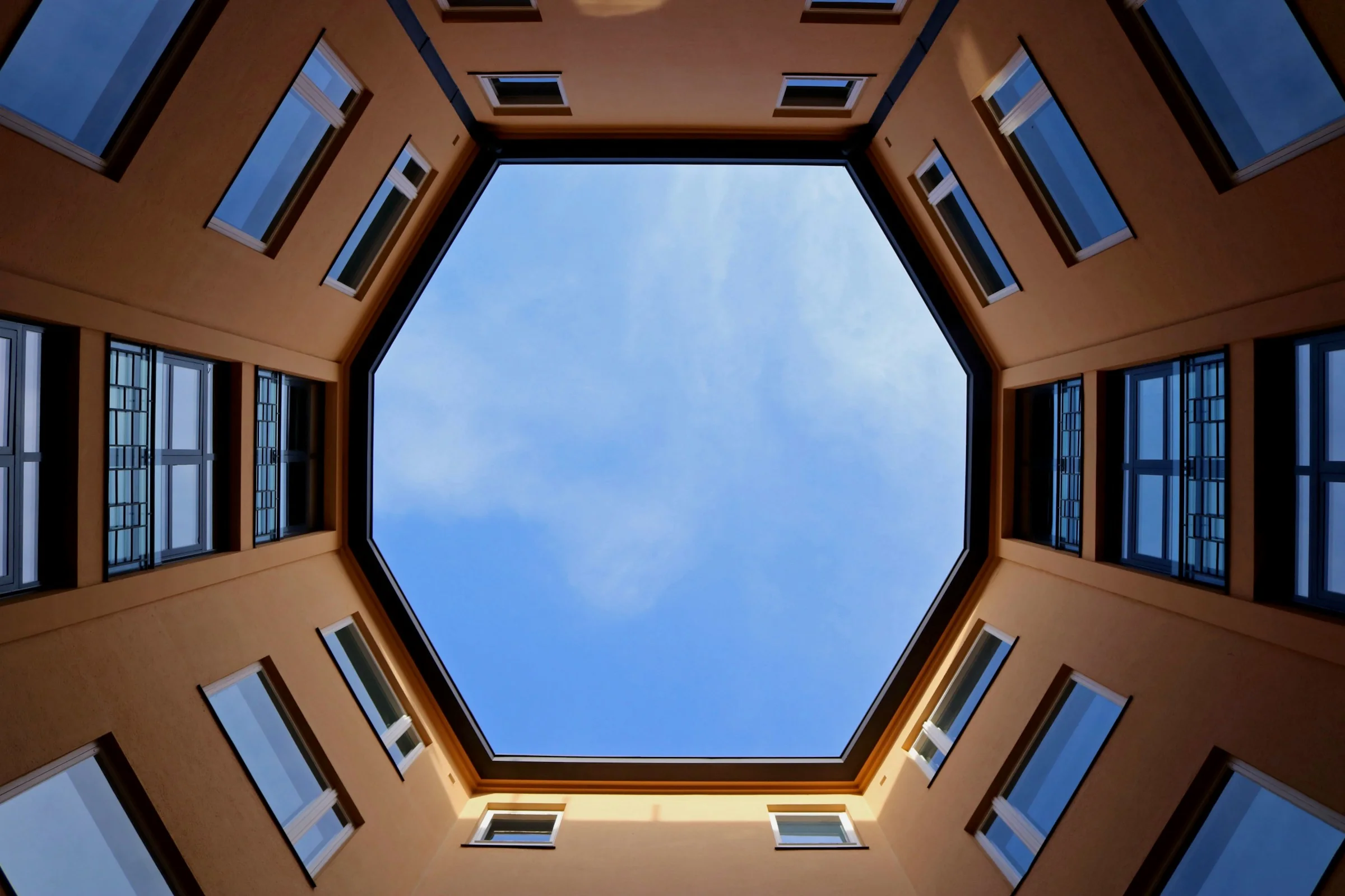 Looking straight up from an octagonal courtyard: orange walls and rows of windows frame a blue sky.